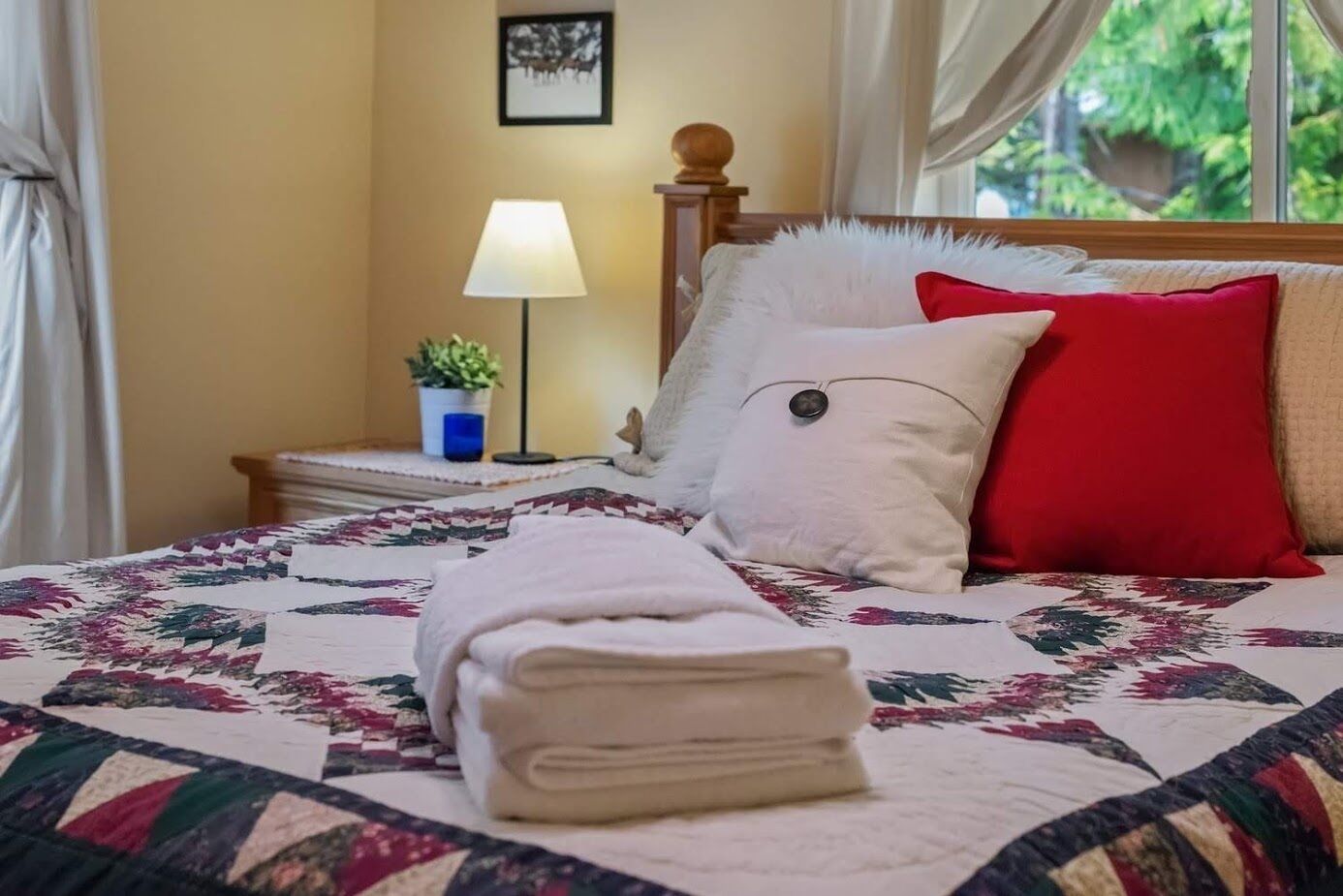 A neatly made bed with a quilt, a red pillow, and a white pillow. There is a small table with a lamp and a plant beside the bed.
