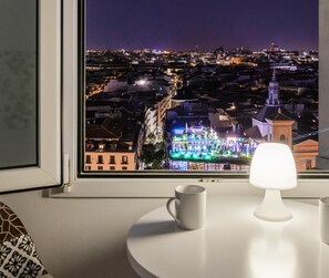 Vista desde una ventana que domina un paisaje urbano nocturno, con edificios iluminados y un campanario de iglesia destacada.