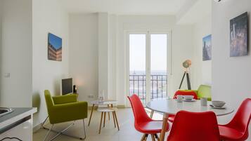 Un salon moderne avec un coin repas, comprenant une grande fenêtre offrant une vue sur la ville, un téléviseur et un lampadaire sur trépied.