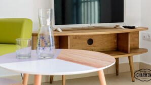 En moderne stue med et fladskærms-TV på et træstel, en grøn sofa, et lille hvidt bord med en vase i glas og en fjernbetjening.