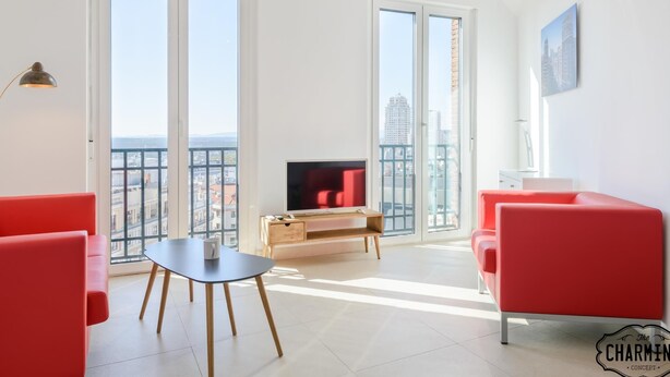 Un salon moderne avec un téléviseur, des fauteuils rouges, une petite table et de grandes fenêtres donnant sur la ville.