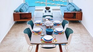 Un espace repas avec une grande table dressée pour six personnes, donnant sur une marina où sont amarrés des bateaux et sur une île tropicale.