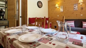 A dining area with a table set for four, a wooden wall with wall-mounted lamps, and a sofa in the background. - Le Portail 27 - Cosy 2-room apartment ski-in ski-out (LES AVANCHERS VALMOREL)