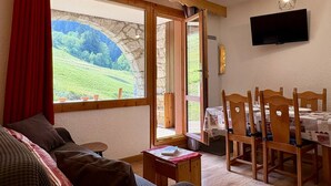 A cozy living room with a sofa, a dining table with chairs, a TV mounted on the wall, and a large window with a view of greenery. - Le Portail 27 - Cosy 2-room apartment ski-in ski-out (LES AVANCHERS VALMOREL)