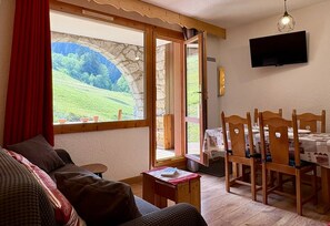A cozy living room with a sofa, a dining table with chairs, a TV mounted on the wall, and a large window with a view of greenery. - Le Portail 27 - Cosy 2-room apartment ski-in ski-out (LES AVANCHERS VALMOREL)