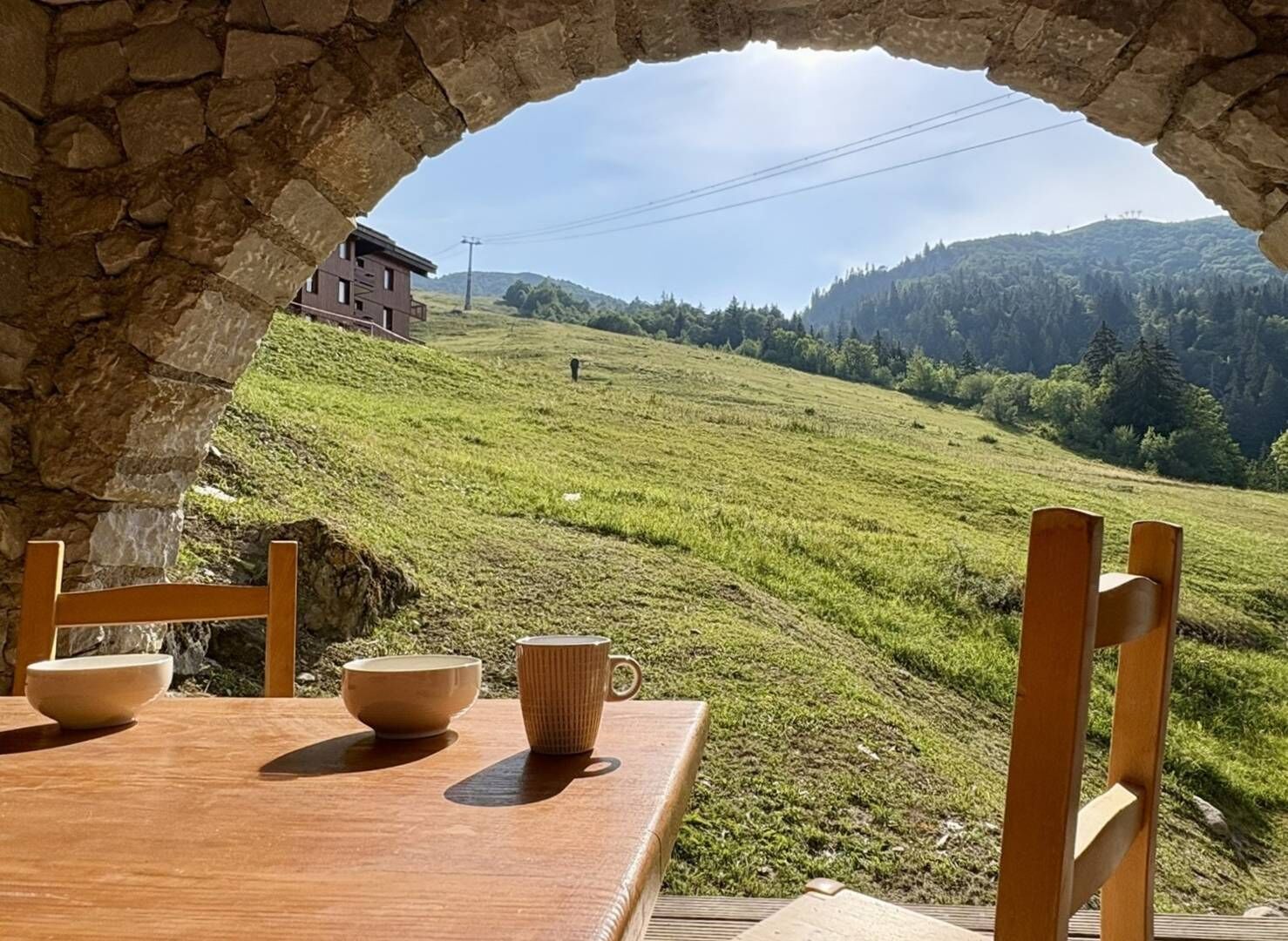 Une table en bois, entourée de chaises et de bols, placée devant une colline verdoyante et un arc en pierre.