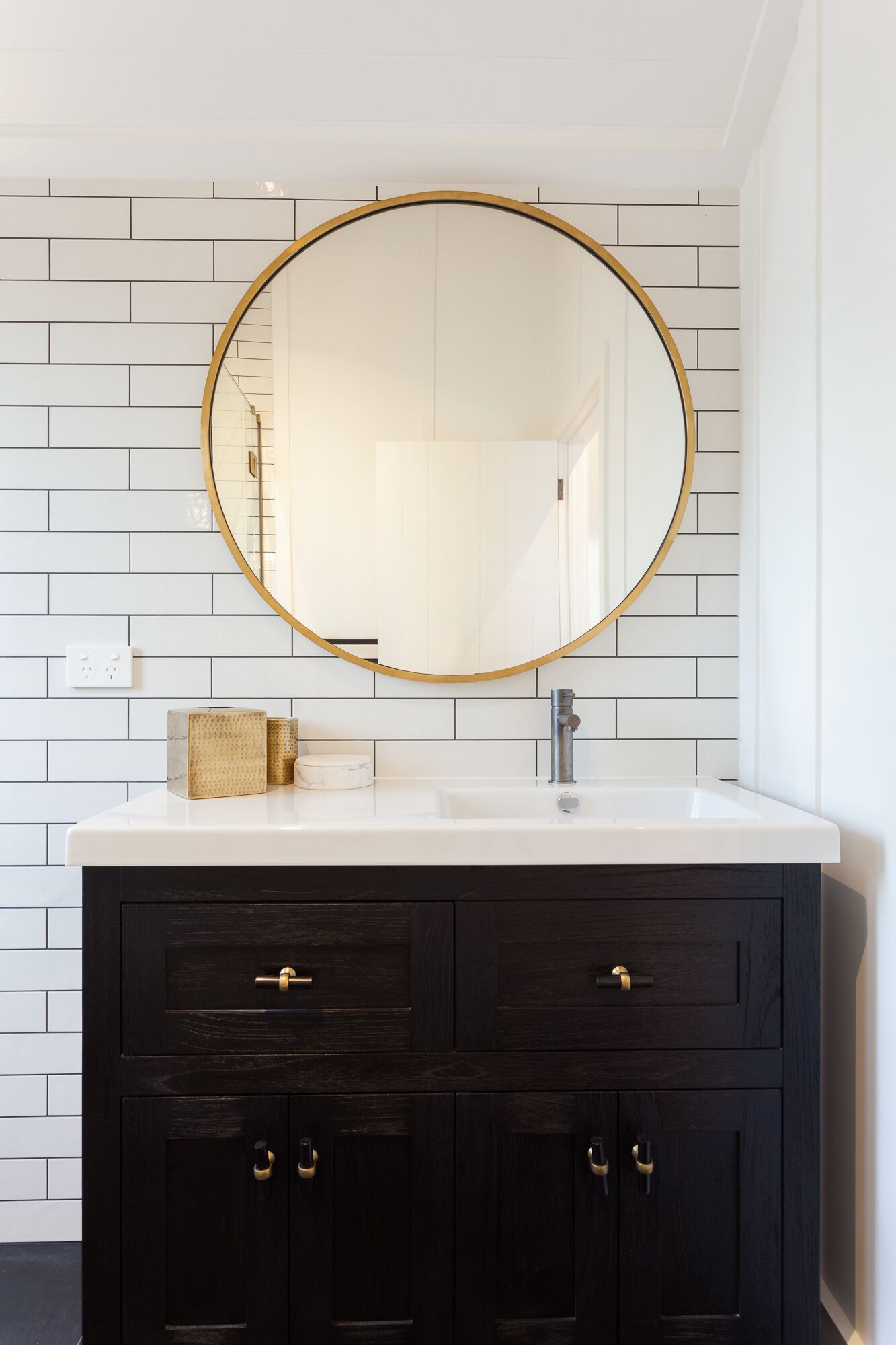 A bathroom with a white tiled wall, a white sink, a dark wooden vanity with gold handles, and a gold-framed mirror.
