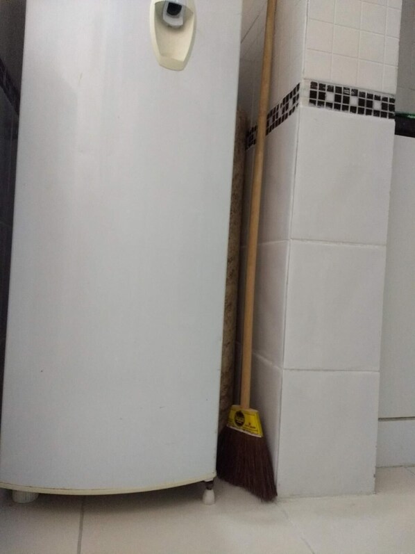 Un refrigerador blanco con dispensador de hielo incorporado, una escoba apoyada en él y una pared de azulejos con un diseño blanco y negro.