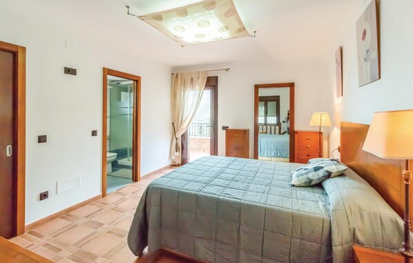 Une chambre à coucher comprenant un lit, une table de chevet, une lampe, un luminaire au plafond et une porte donnant sur une salle de bain.