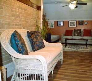 A wicker sofa with patterned cushions, a white armchair, and a wooden coffee table in a room with a brick wall and wooden flooring.