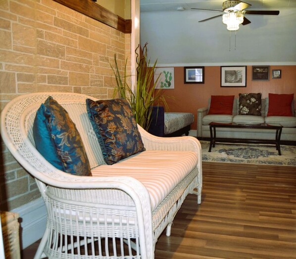 A wicker sofa with patterned cushions, a white armchair, and a wooden coffee table in a room with a brick wall and wooden flooring.