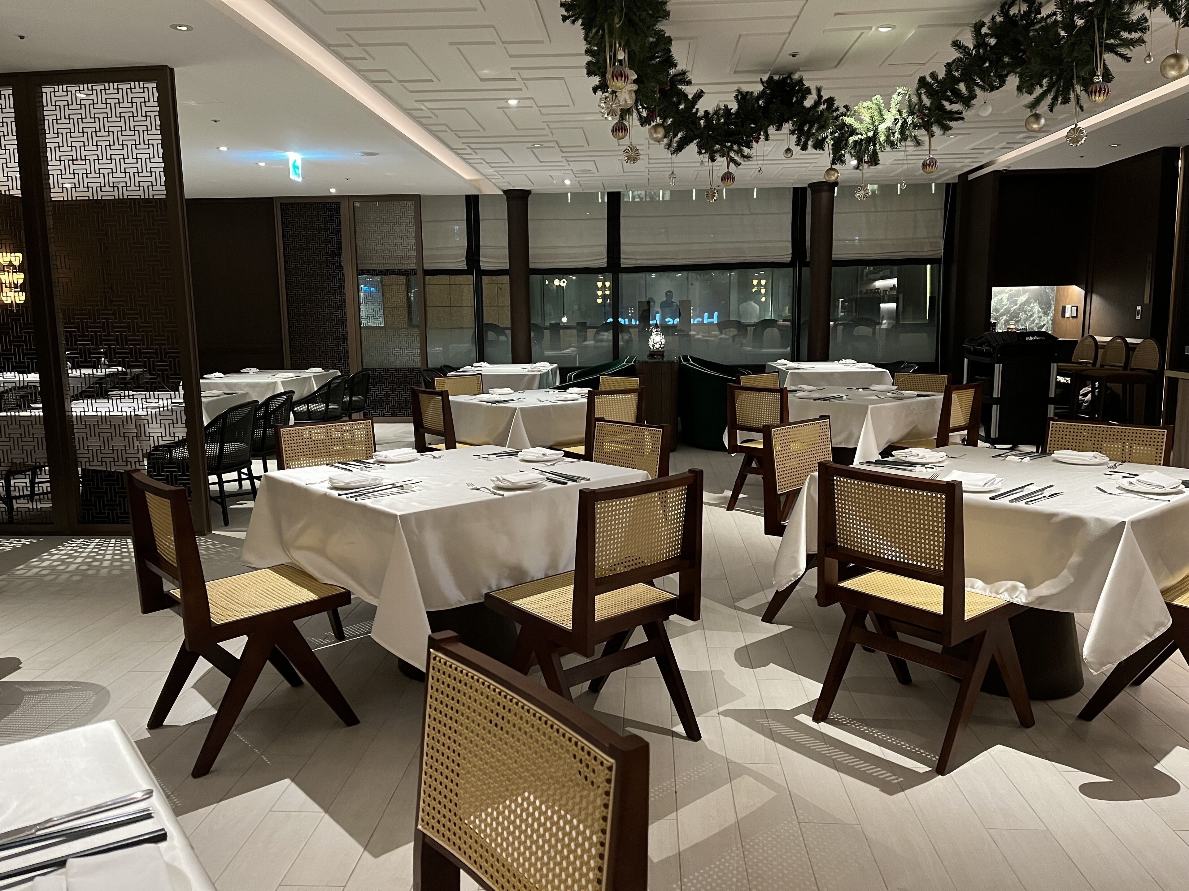 A restaurant with multiple tables set for dining, featuring a modern interior with wooden chairs and a ceiling decorated with greenery.