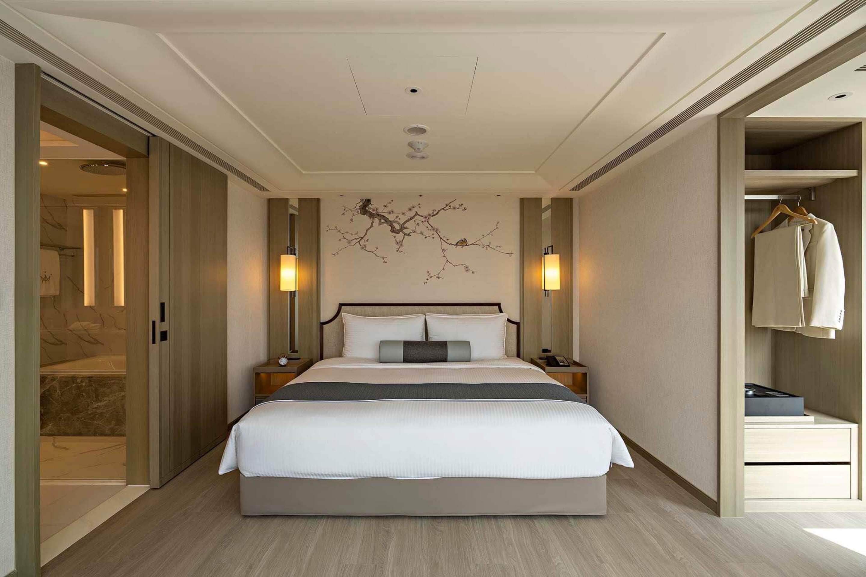 A modern hotel room with a large bed, bedside tables, a wardrobe, and a wall-mounted artwork.