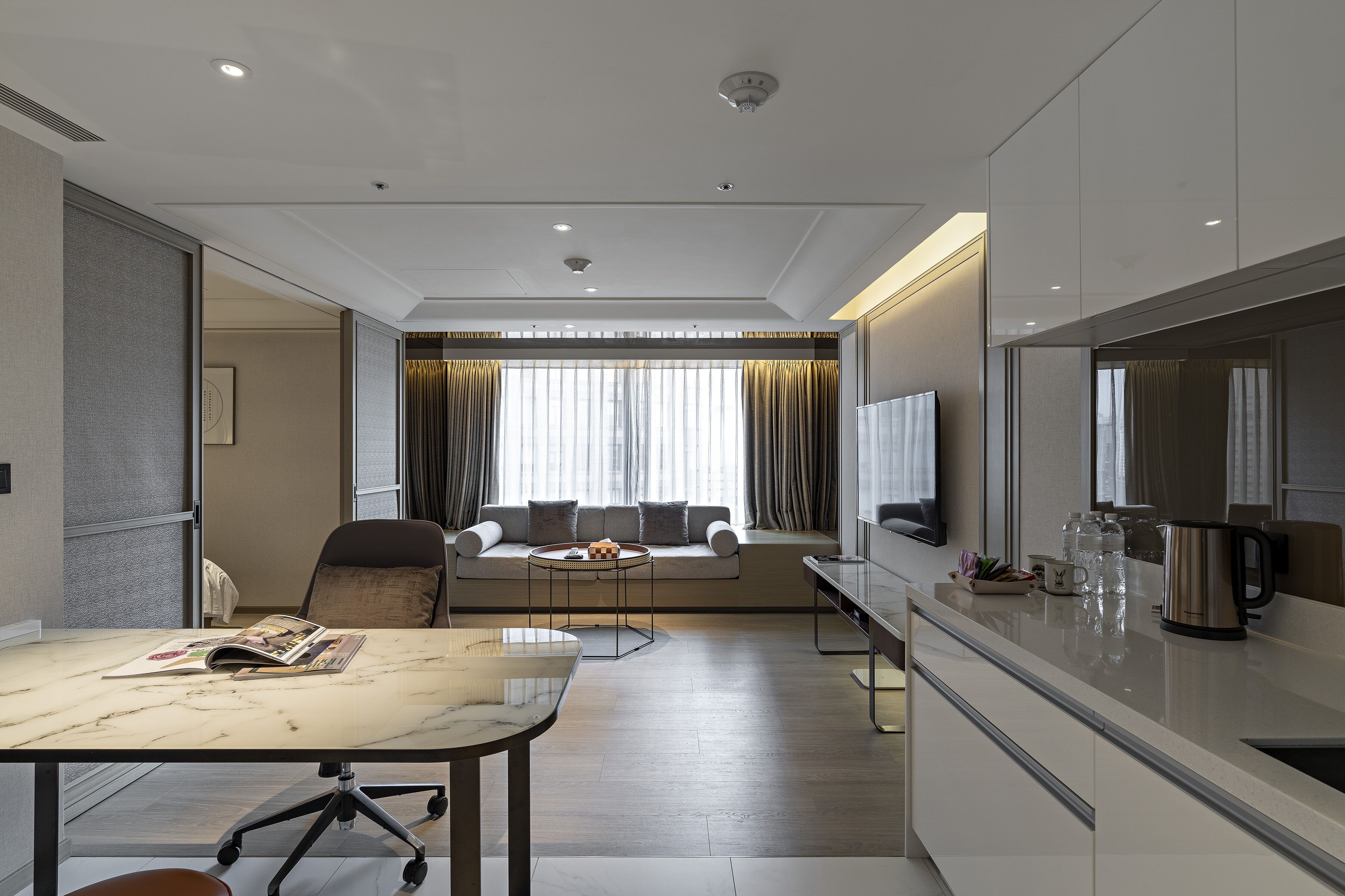 A modern hotel room with a marble dining table, a sofa with cushions, a TV, and a kitchen area with a kettle and glasses.