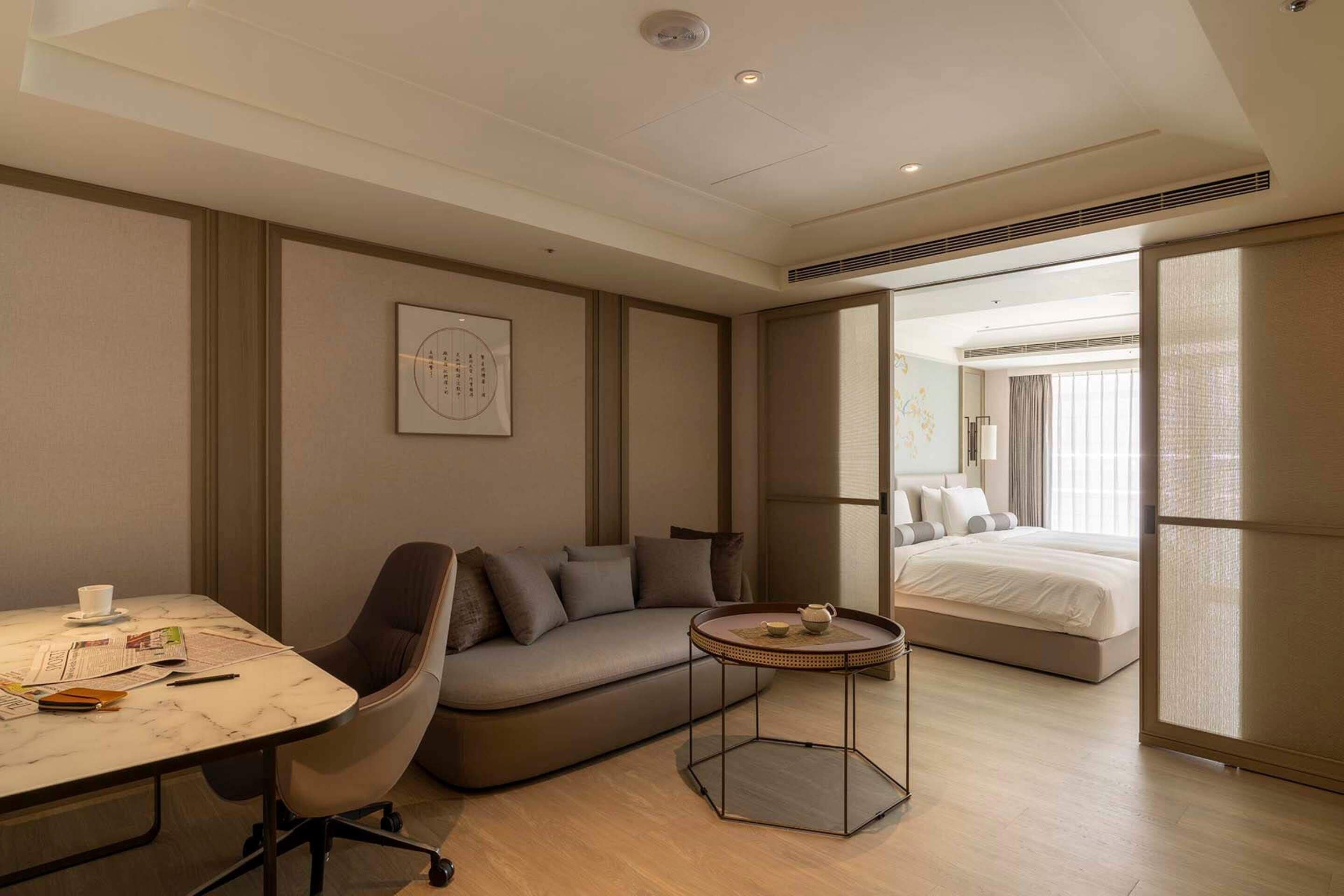A modern hotel room with a sofa, a small round table, a bed, and a large window.