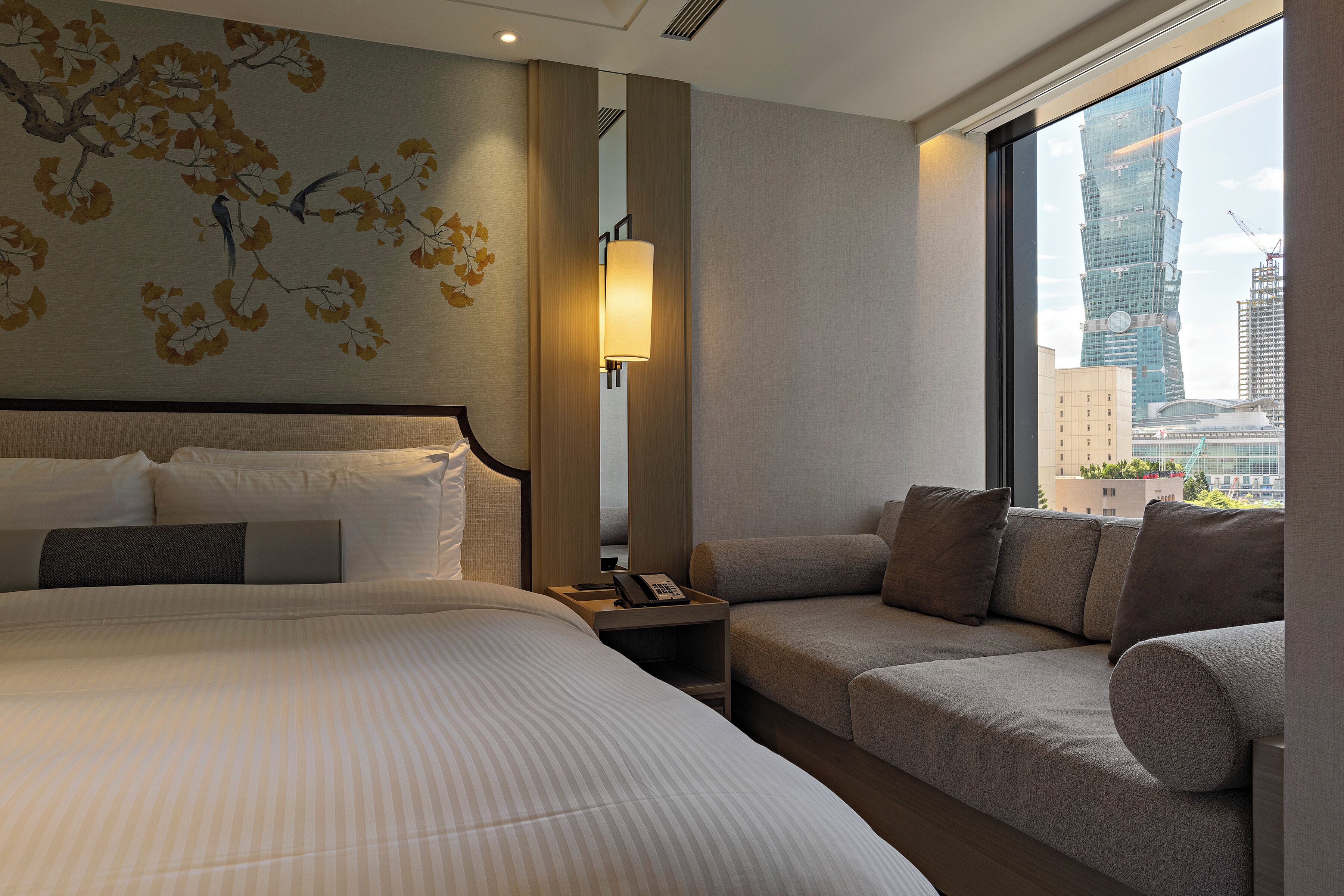 A hotel room with a large bed, a sofa, a nightstand with a lamp, and a view of a cityscape.