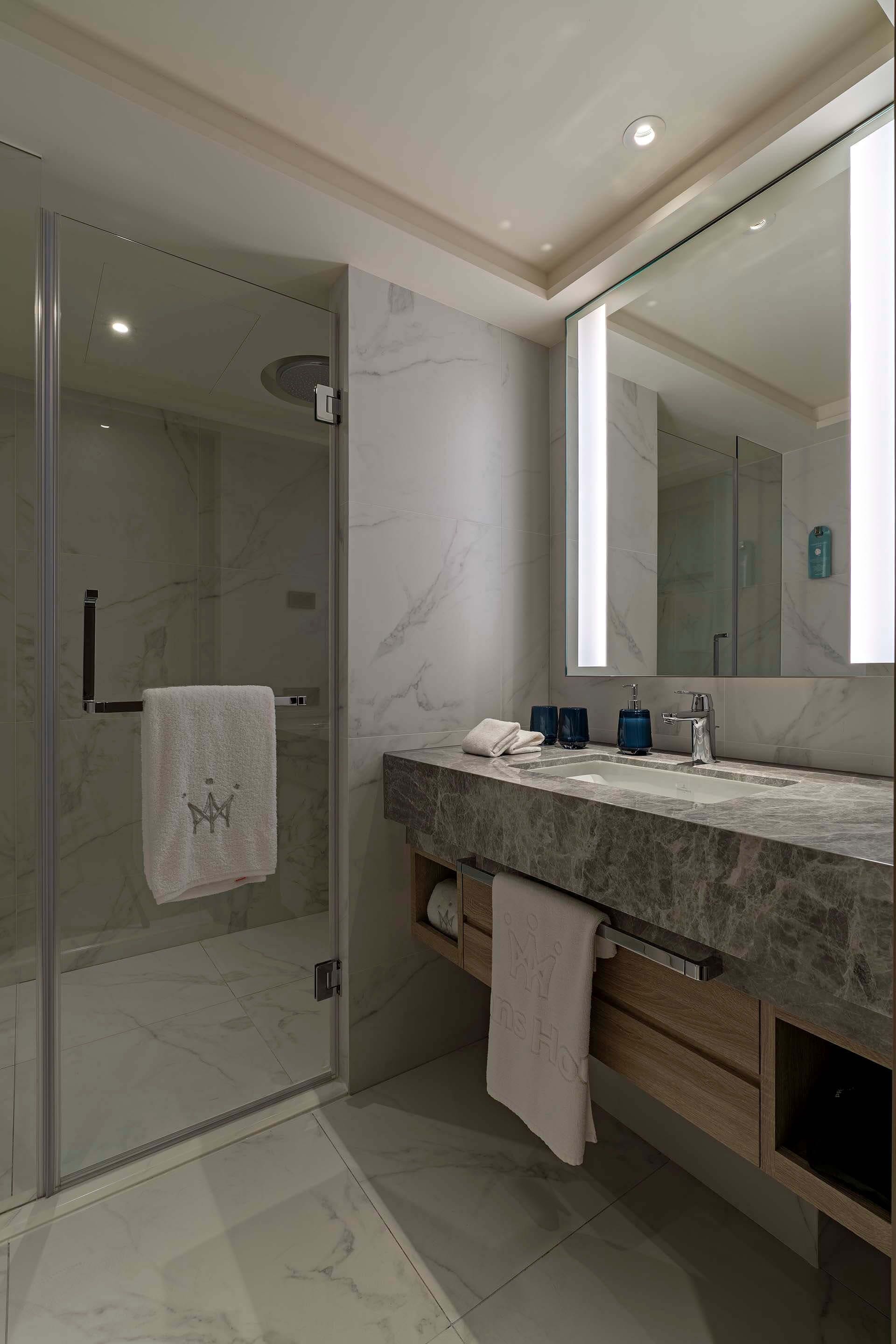 A modern bathroom with a glass shower enclosure, a marble-tiled floor, a large mirror, and a marble countertop with a sink and towel rack.