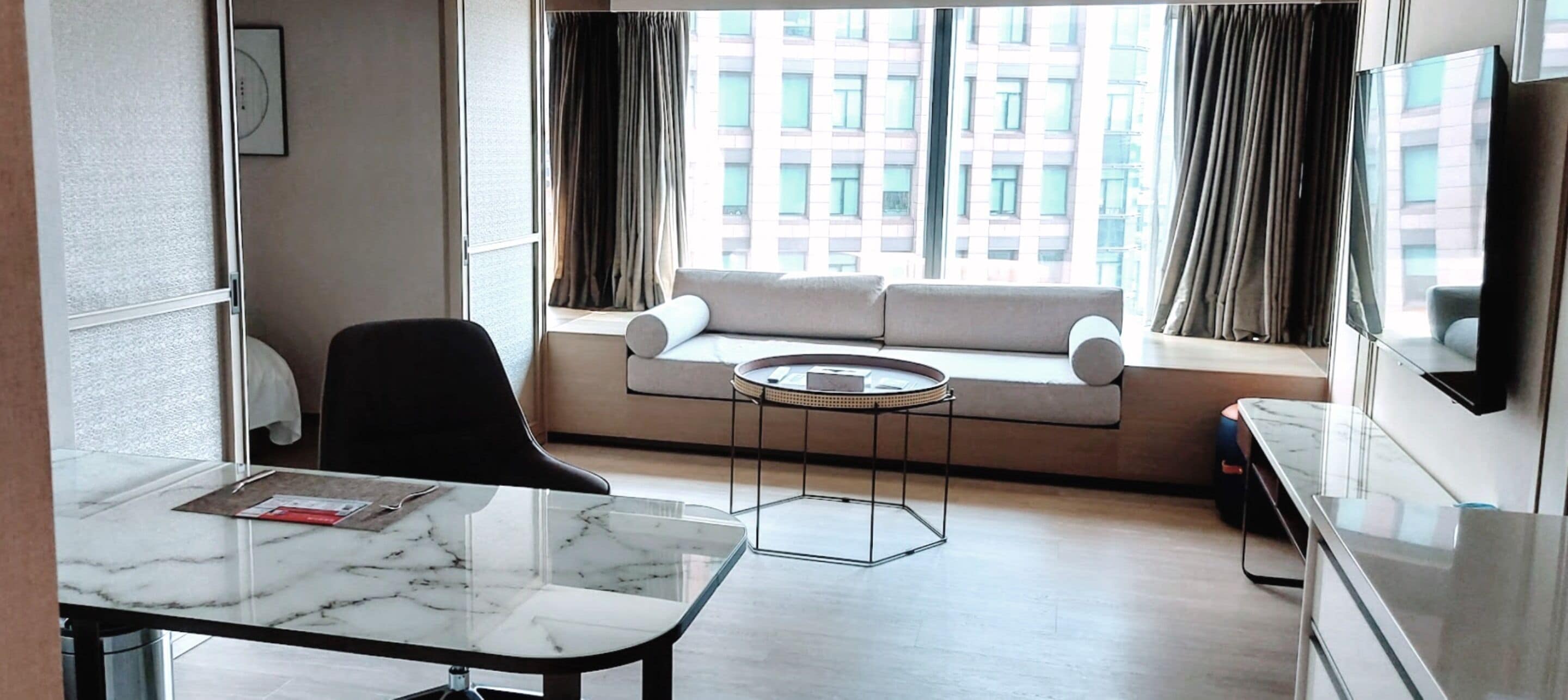 A modern hotel room with a sofa by a large window, a round coffee table, a black chair, and a marble-topped desk.