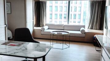 A modern hotel room with a sofa by a large window, a round coffee table, a black chair, and a marble-topped desk.