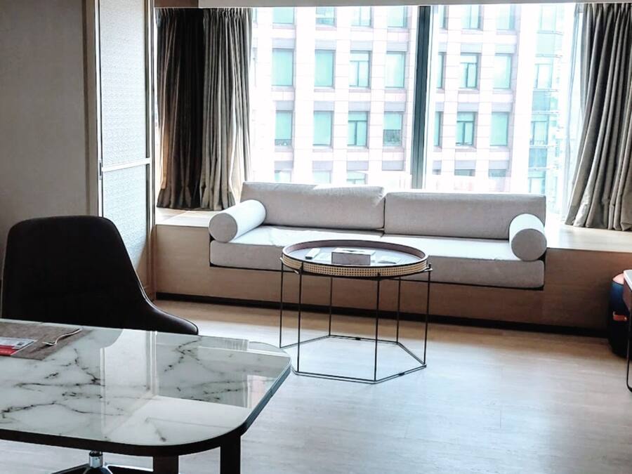 A modern hotel room with a sofa by a large window, a round coffee table, a black chair, and a marble-topped desk.