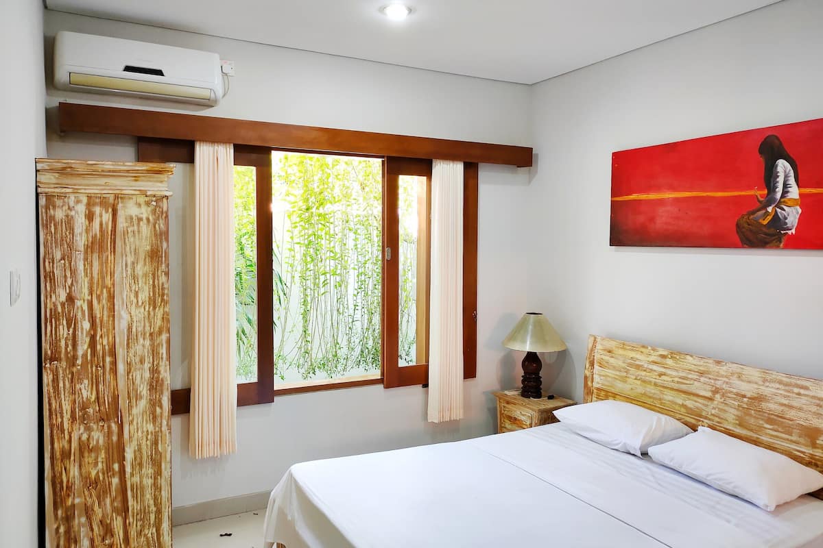 A bedroom with a bed, a nightstand with a lamp, a painting, and a window with curtains.