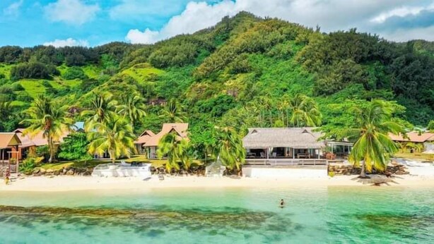 Un hotel sulla spiaggia con bungalow sull'acqua, acque turchesi cristalline e dolci colline verdi sullo sfondo.