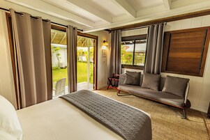 A bedroom with a bed, a sofa, and a chair. There is a window with curtains, a door, and a view of a garden.