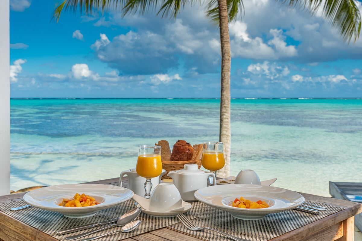 A breakfast setup with plates of food, two glasses of orange juice, a basket of bread, and a white teapot on a table with a view of the ocean and palm trees.