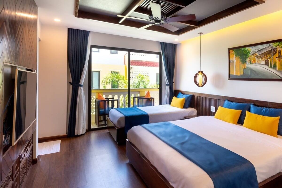A hotel room with two beds, a ceiling fan, a large window with curtains, a painting on the wall, and a balcony with a table and chairs.