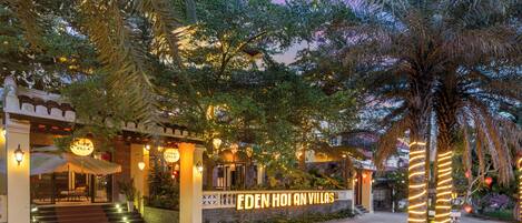 A well-lit entrance to Eden Hoi An Villas with a clear pathway leading to the main building, surrounded by lush greenery and palm trees.