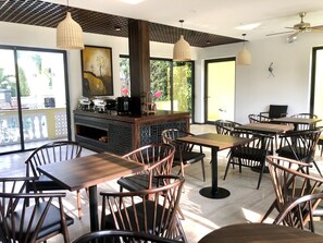 A restaurant with wooden tables and chairs, a central counter with a coffee machine, and large windows allowing natural light.