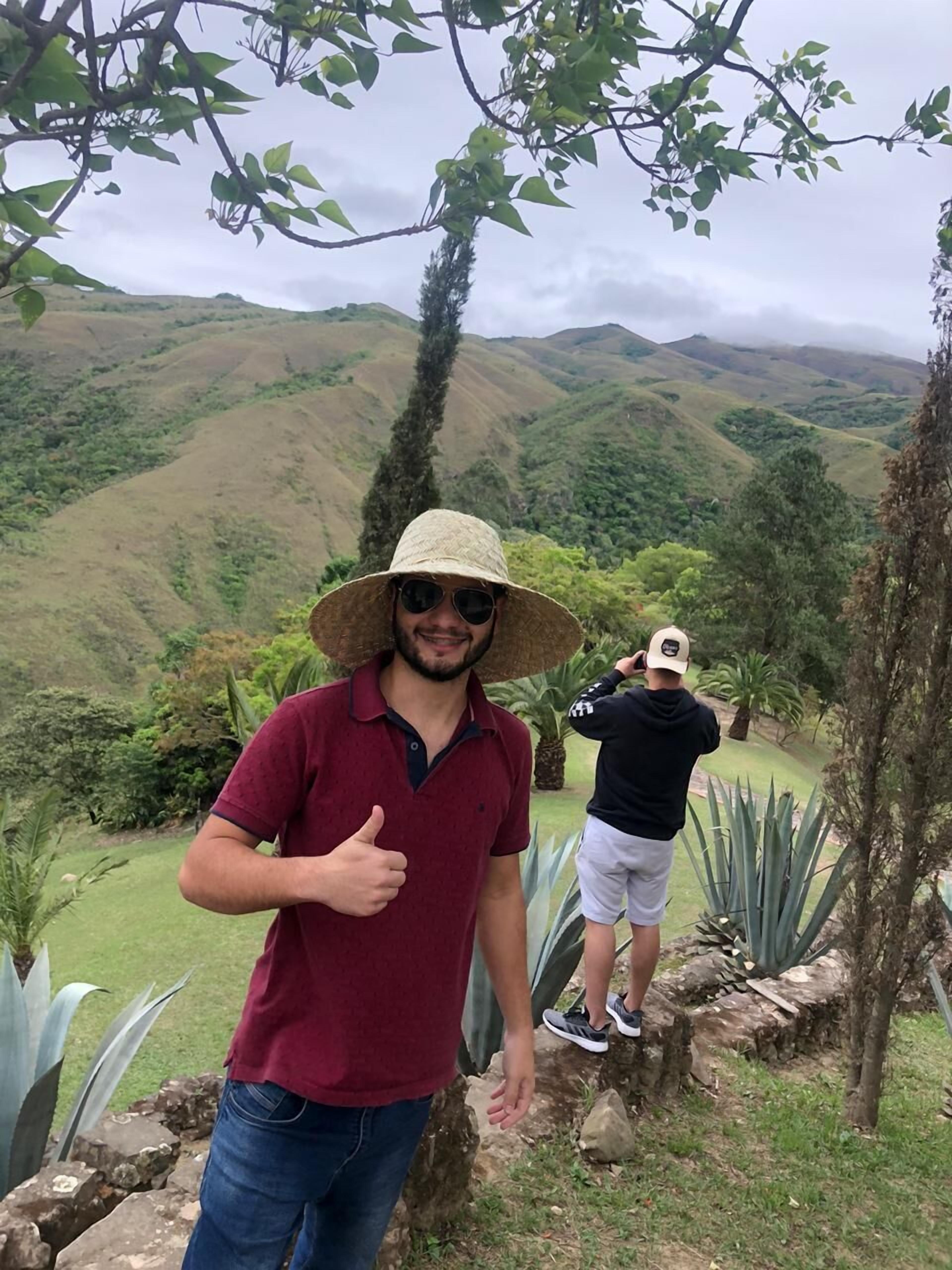 Een persoon in een rood shirt en strohoed die een duimpje omhoog steekt, met een bergachtig landschap op de achtergrond.