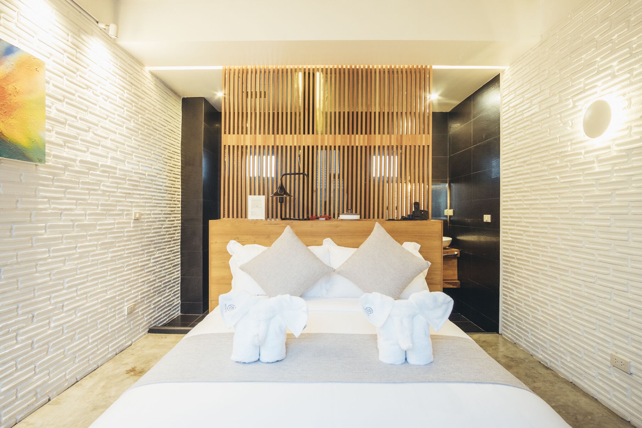 A modern hotel room with a large bed, two white elephant-shaped pillows, a wooden headboard, and a decorative wooden panel above the bed.