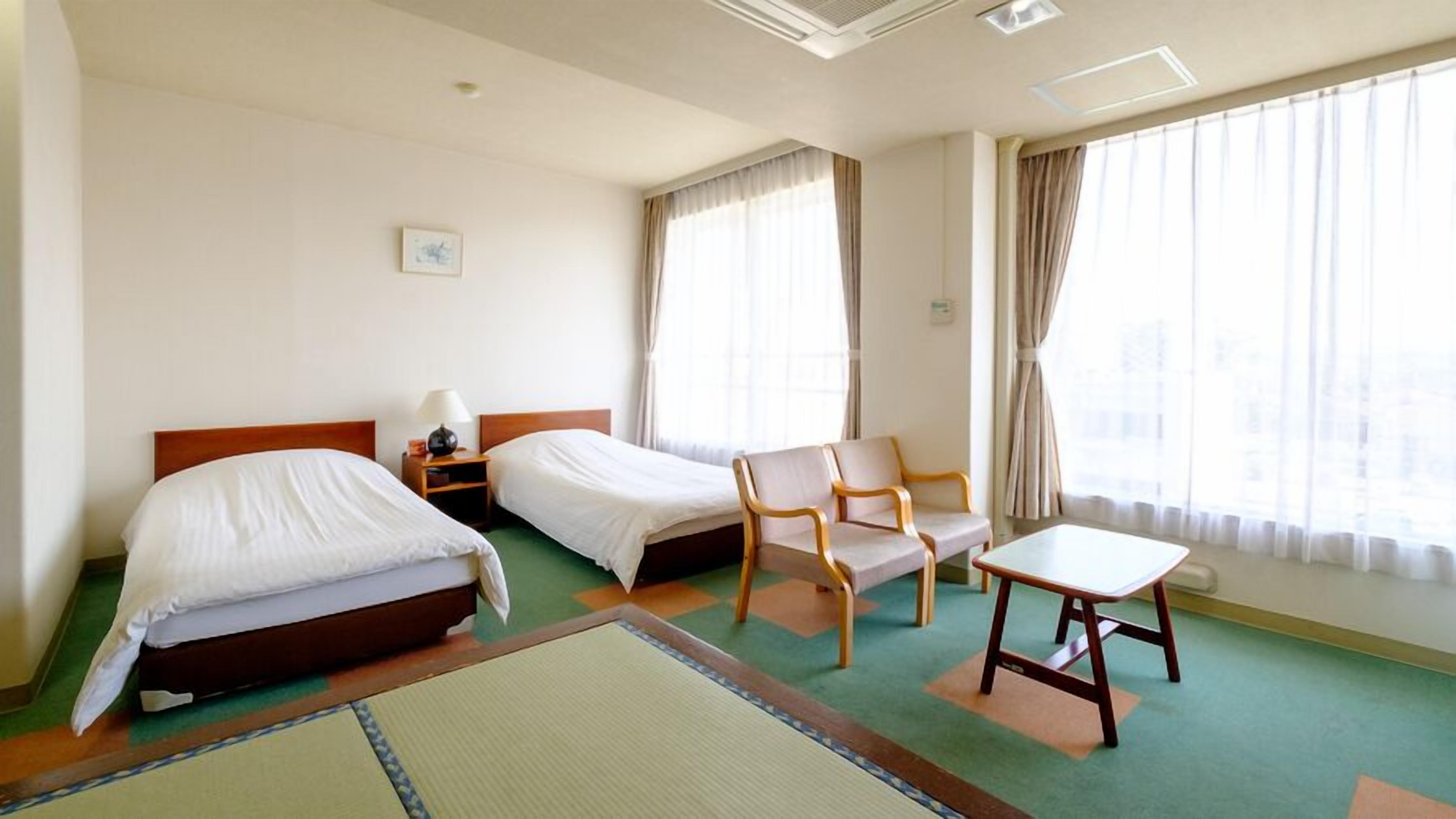 A hotel room with two beds, a chair, a table, and large windows with curtains.