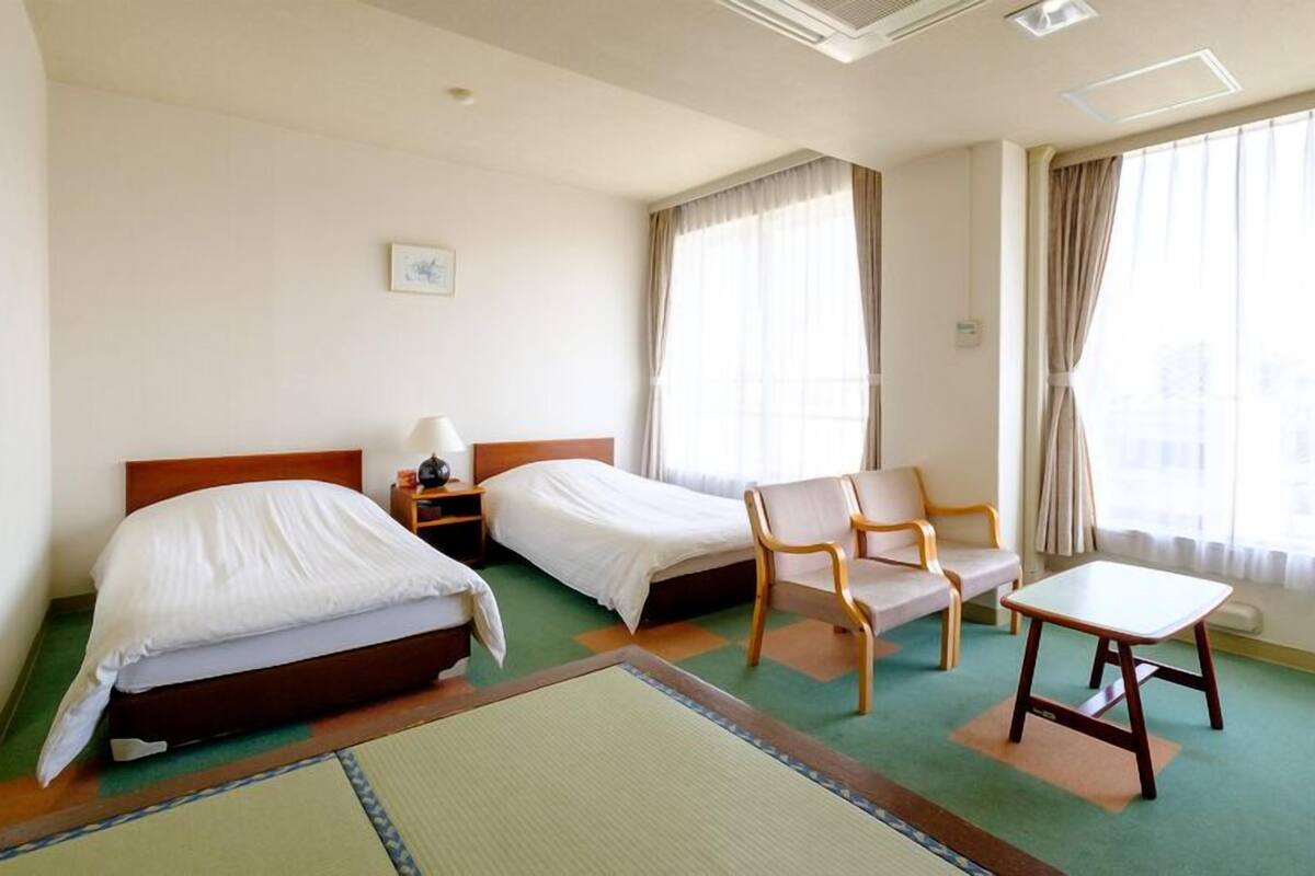 A hotel room with two beds, a chair, a table, and large windows with curtains.