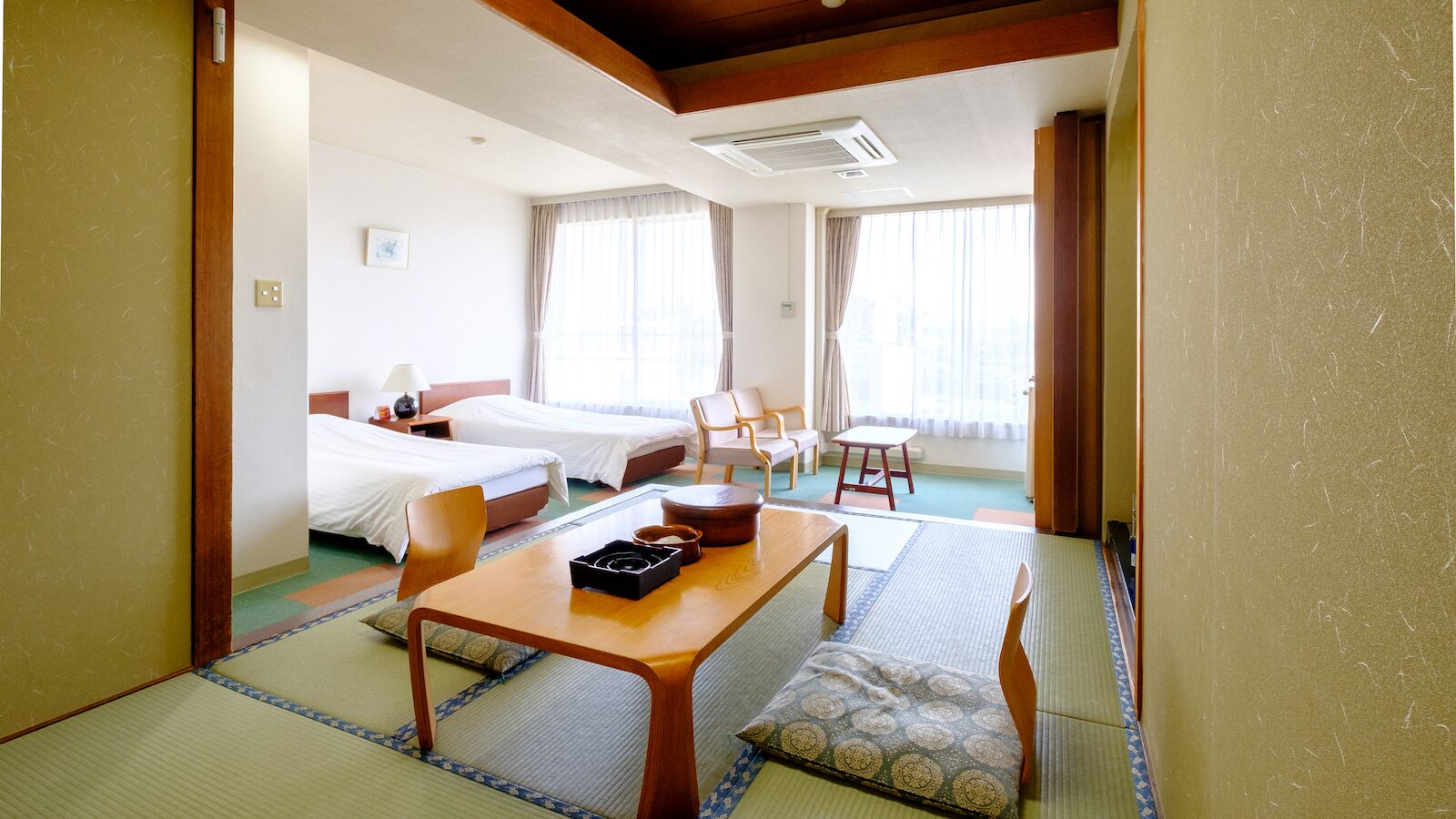 A hotel room with two beds, a wooden table, chairs, a small table, and a view of the city through large windows.