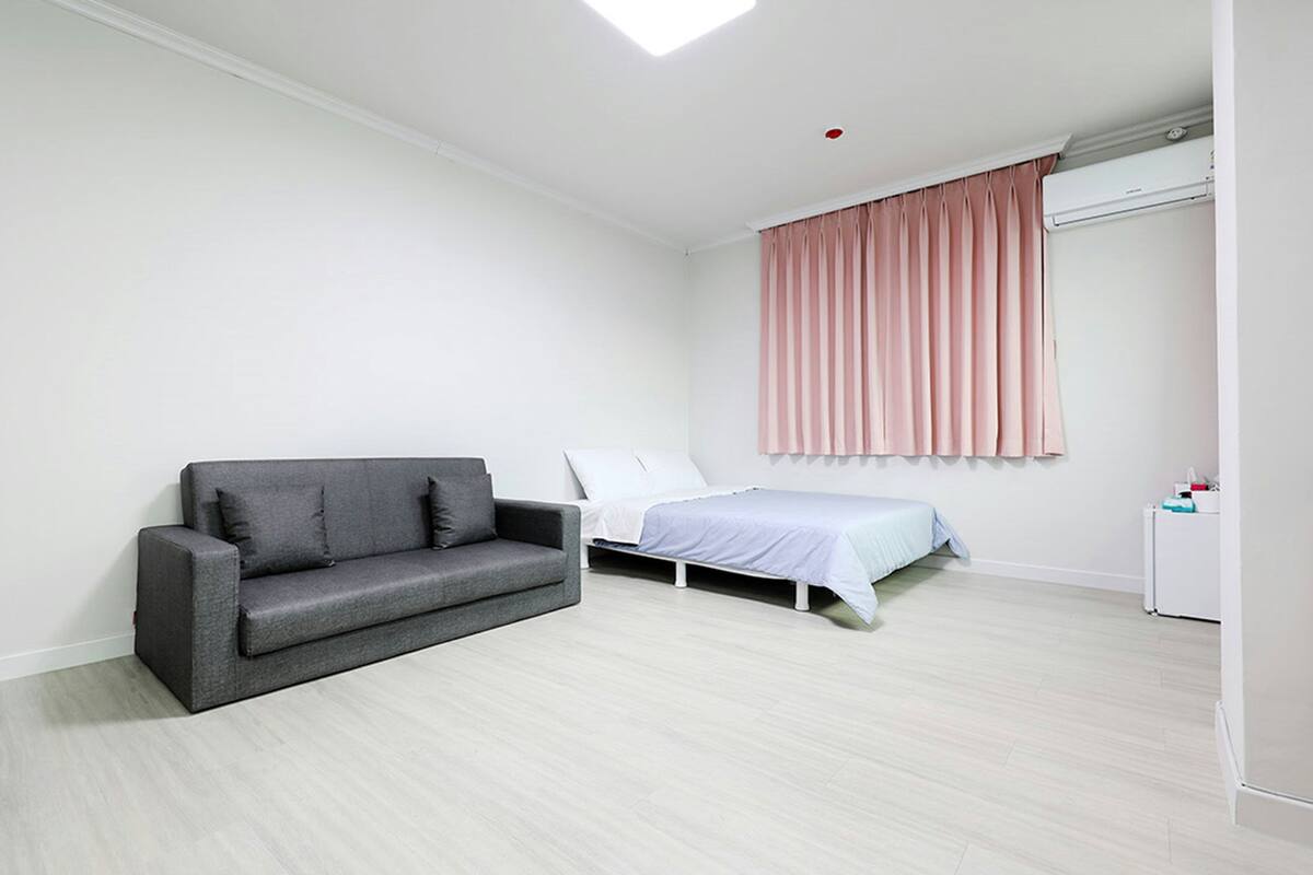 Ein modernes Zimmer mit einem grauen Sofa, einem Bett mit weißer Matratze und einem rosa Vorhang.