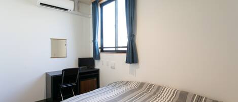 Une chambre avec un lit, un bureau et une chaise. Il y a une fenêtre avec des rideaux, un appareil de climatisation et un miroir au mur.