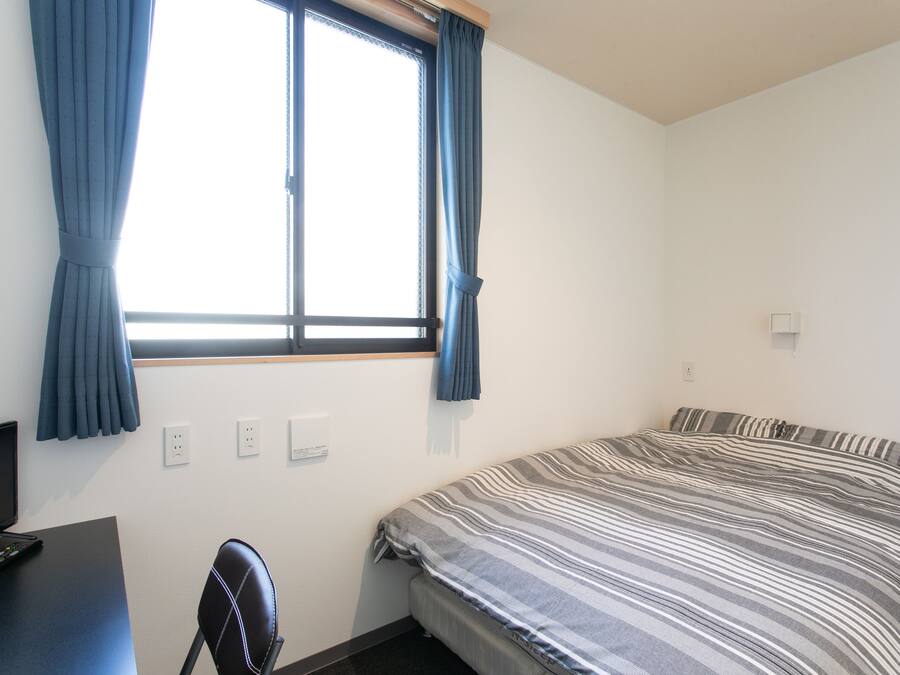 A bedroom with a bed, a desk, a chair, a window with curtains, and a TV.