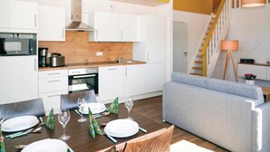 Cozinha moderna com armários brancos, bancada de madeira e forno embutido. Há uma área de jantar com mesa de madeira posta para refeição, e um sofá cinza na sala de estar.