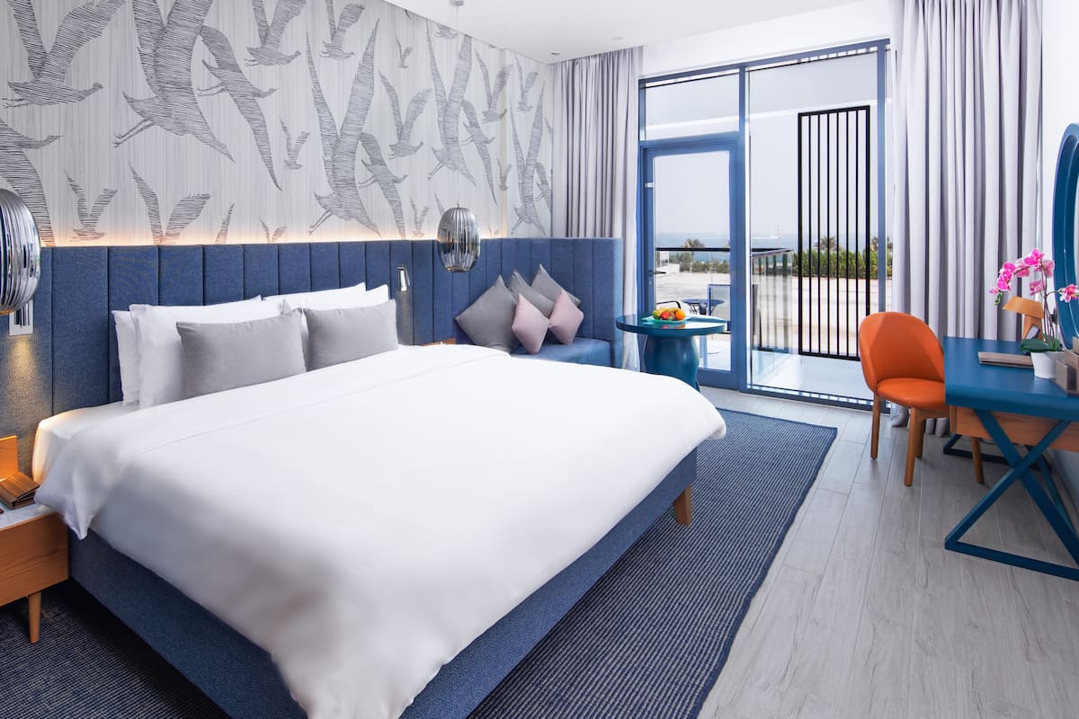 Une chambre d’hôtel moderne avec un grand lit, un bureau avec une chaise, une petite table avec des fruits et une vue sur l’extérieur.