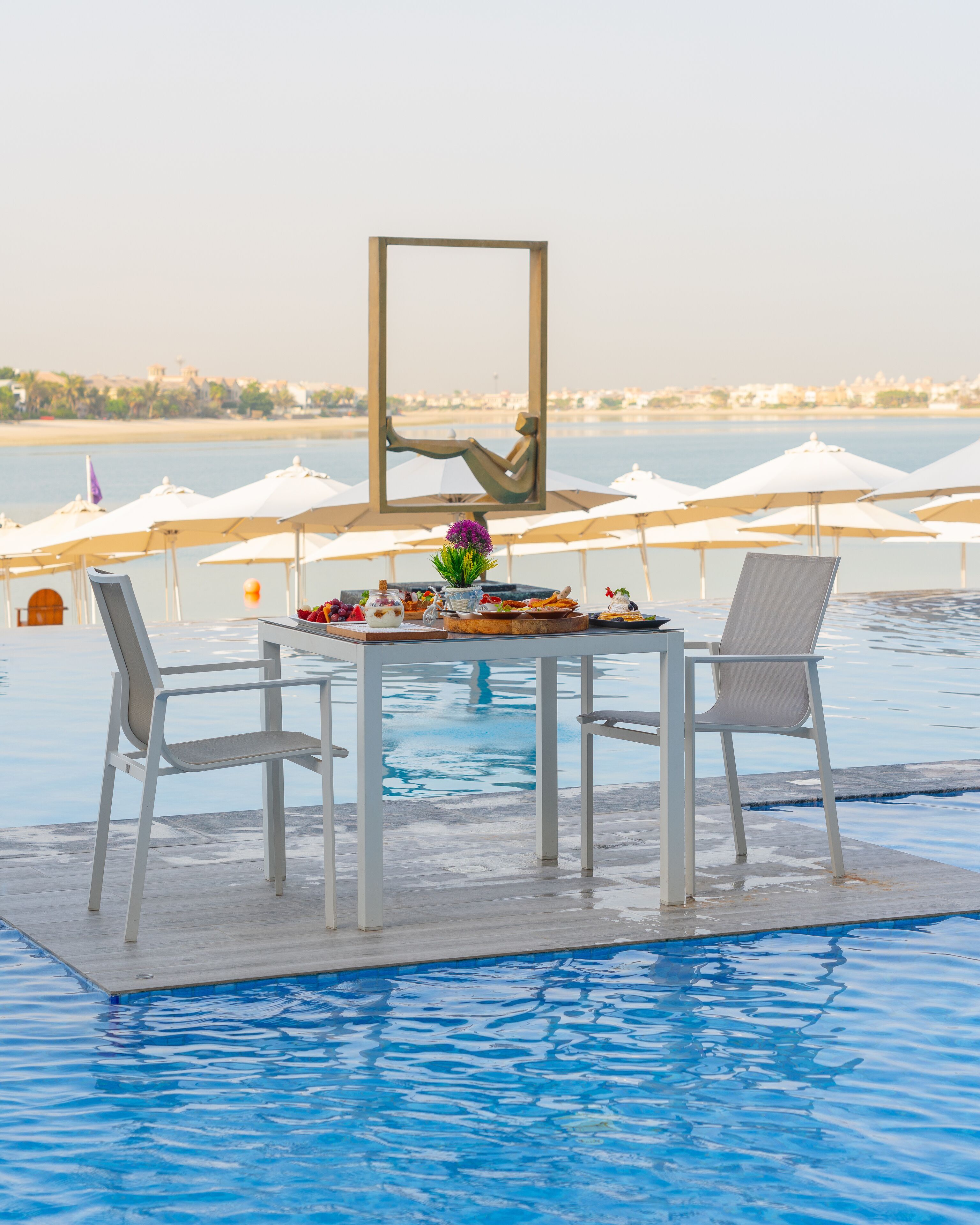 Zona de comedor junto a la piscina con una mesa preparada para dos, con vistas a un cuerpo de agua.