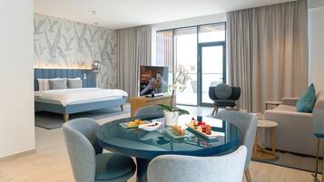 A modern hotel room with a bed, a dining area with chairs and a table, a sofa, and a large window with curtains.