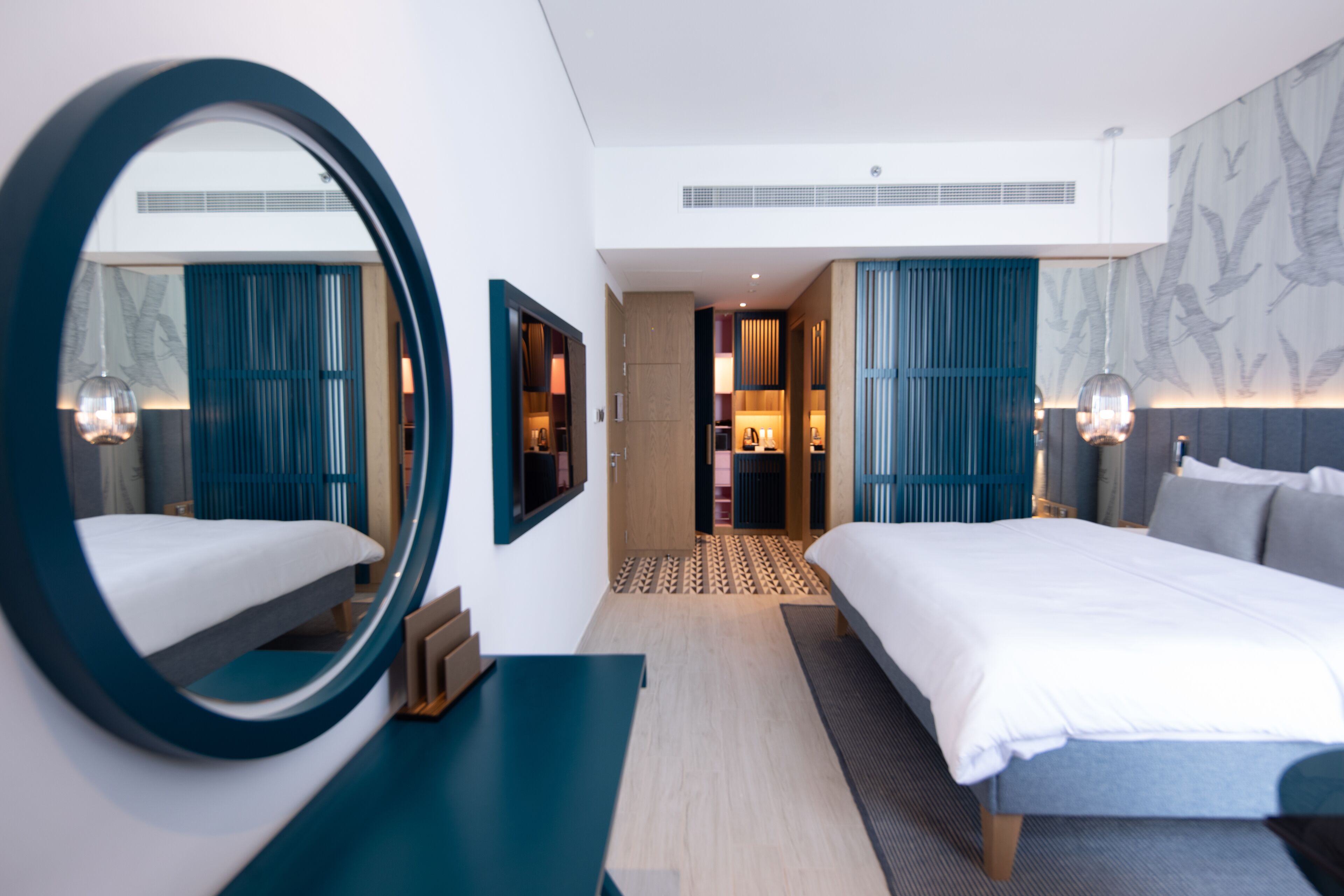 A modern hotel room with a large bed, a round mirror, and a view into the adjacent room.