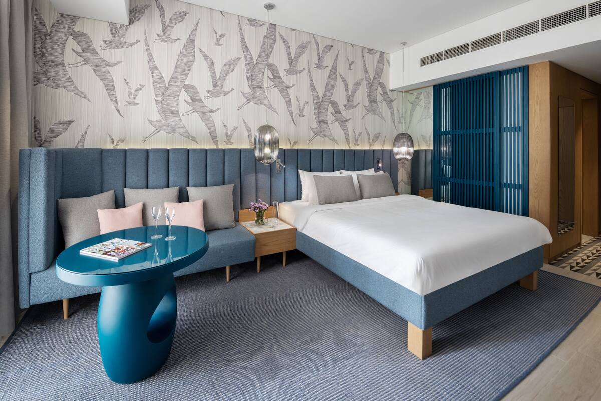 Une chambre d’hôtel moderne dotée d’un grand lit, d’un coin salon avec une table en verre et d’un mur décoratif orné d’un motif représentant des oiseaux.