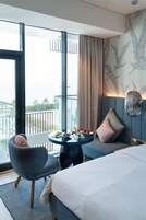A modern hotel room with a large bed, a seating area with a table, and a balcony with a view.