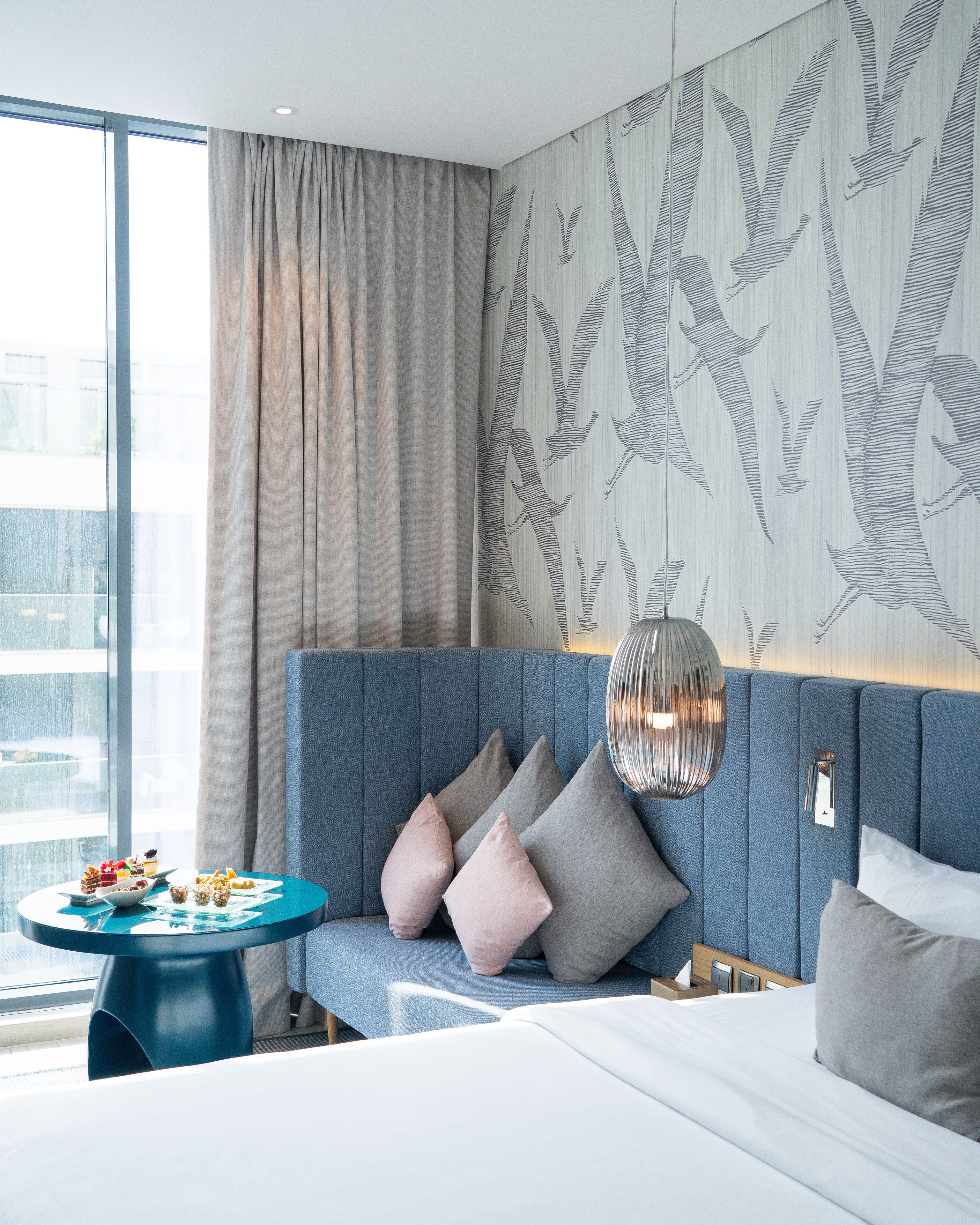 Moderni hotellihuone, jossa on suuri ikkuna, sininen sohva koristetyynyineen, pyöreä pöytä, jossa on tarjolla pientä purtavaa, ja valkoisella patjalla varustettu sänky.