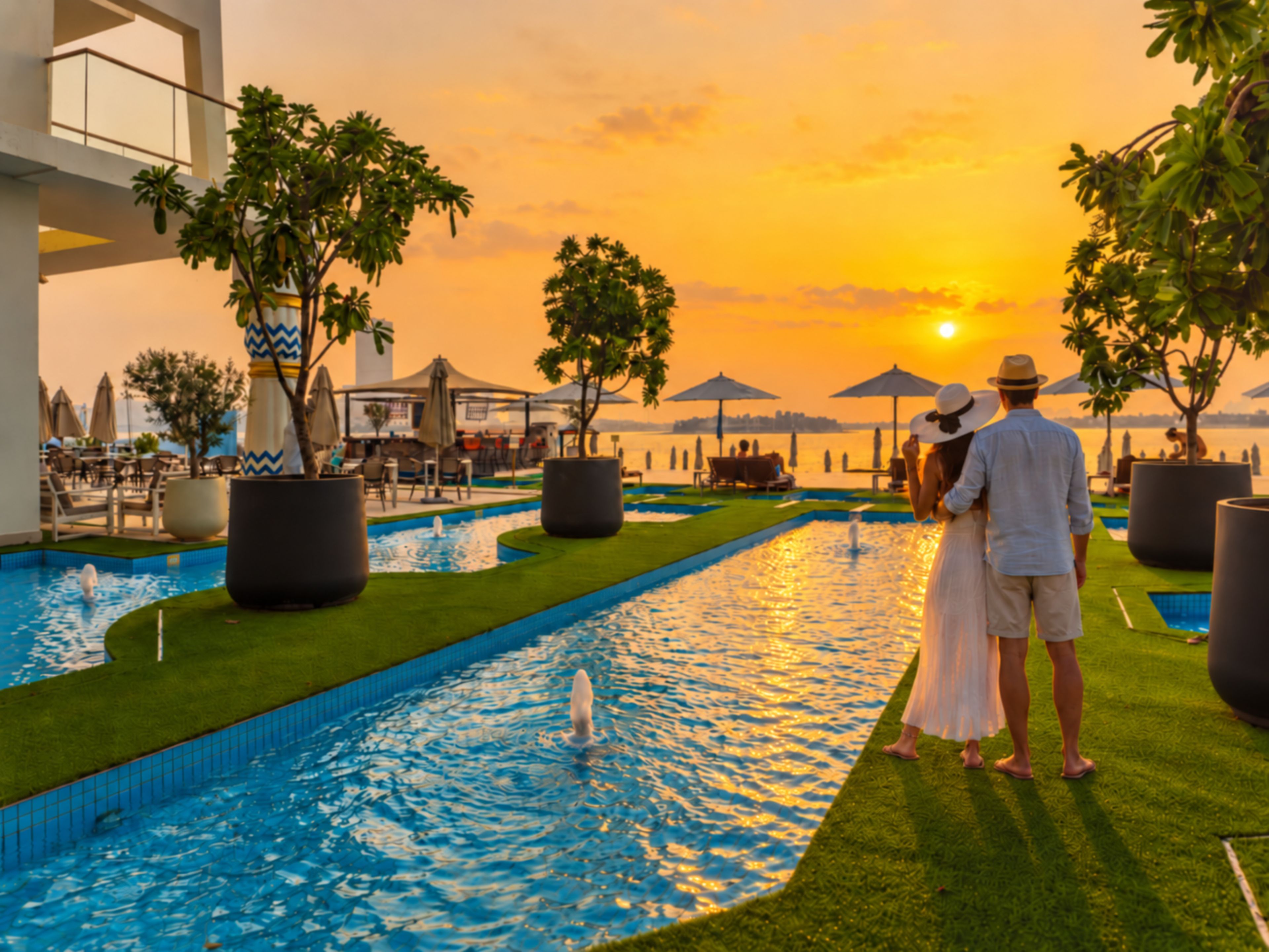 Um casal ao lado de uma piscina com vista para o pôr do sol, cercado por vasos de plantas e guarda-sóis.