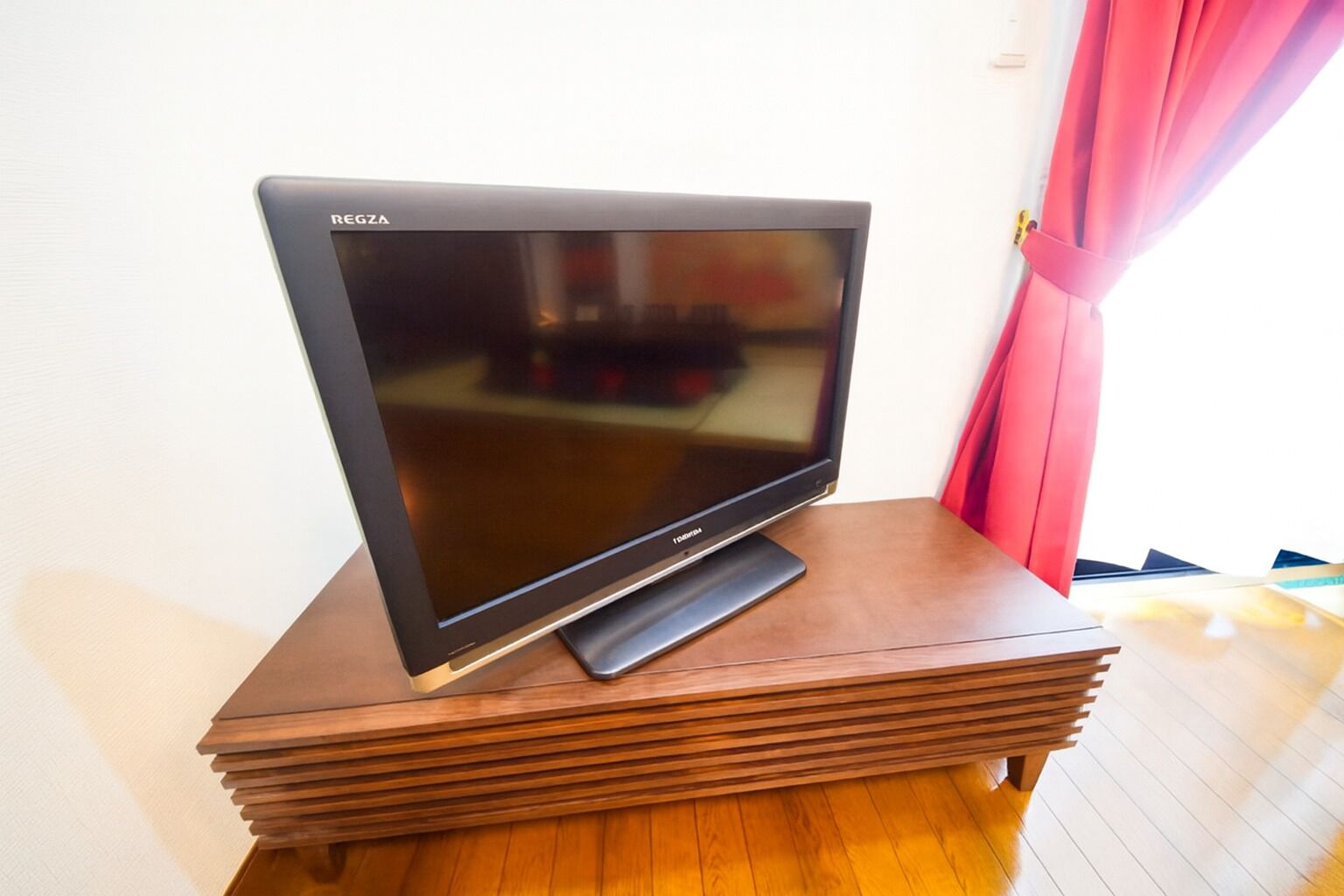 A flat-screen TV on a wooden stand in a room with a white wall and a pink curtain.