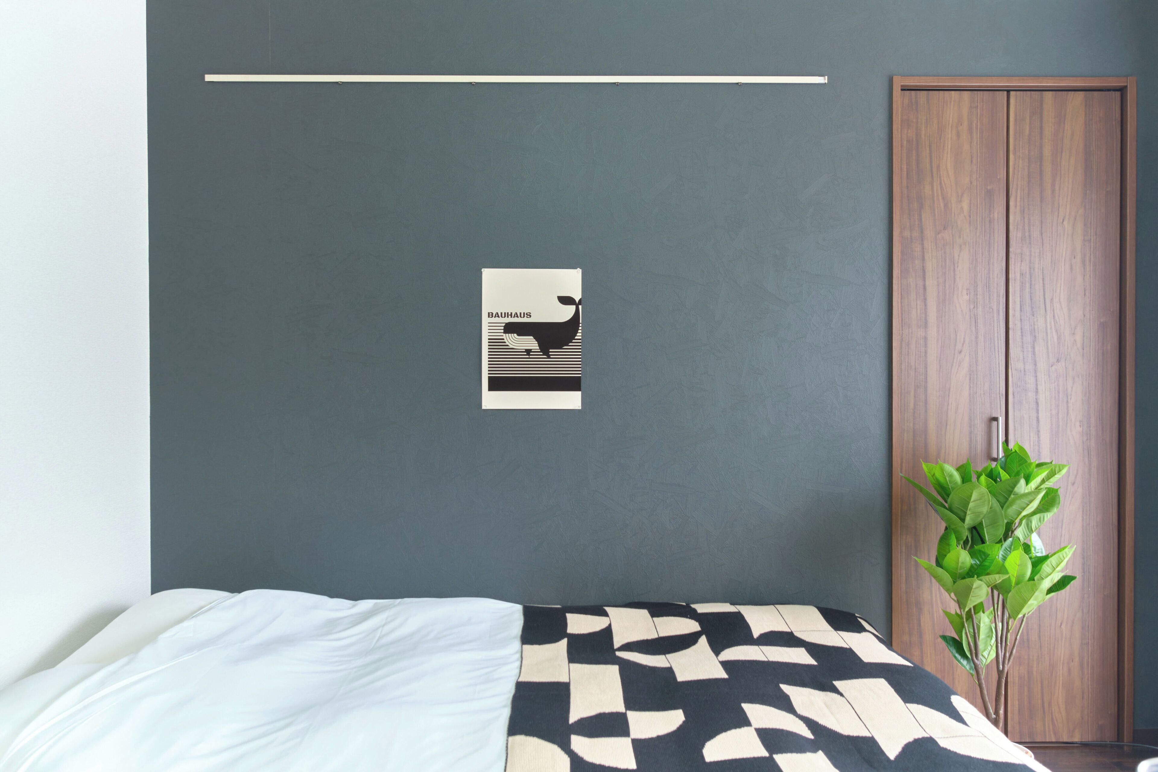 A bedroom with a bed, a wall-mounted poster, a wooden wardrobe, and a potted plant.